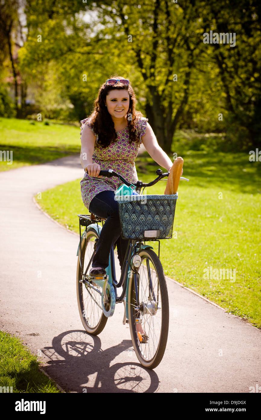 Junge kaukasische Frau, die im Sommer in einem britischen Park auf dem Fahrrad mit einer Flasche und einem Baguette im Korb unterwegs ist Stockfoto