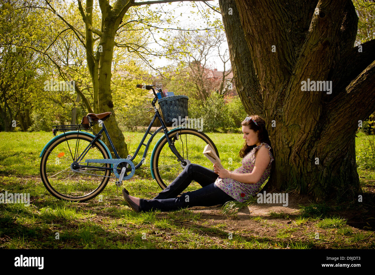 Junge kaukasische Frau sitzt unter einem Baum und entspannt ein Buch zu lesen. Stockfoto