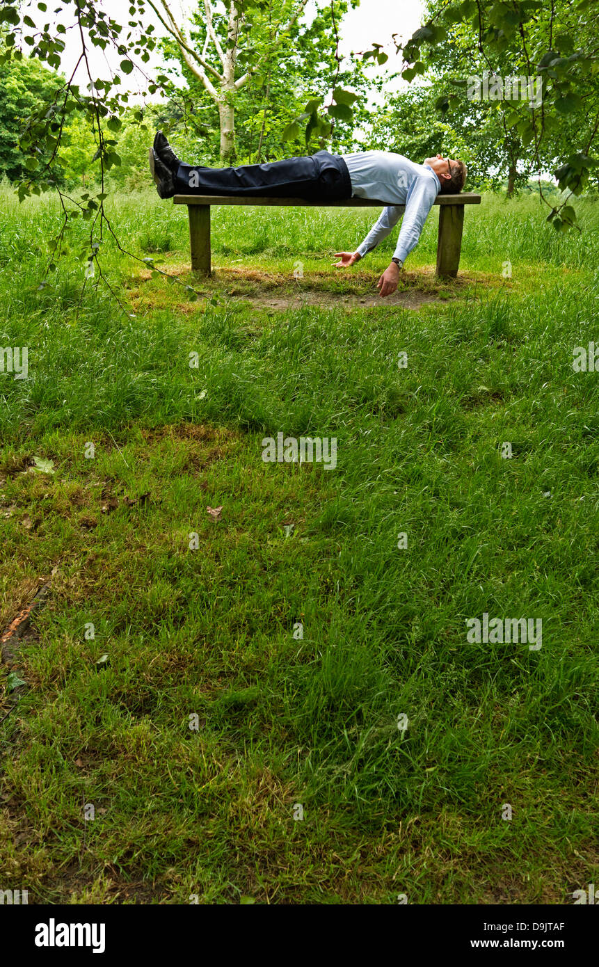 Ein Geschäftsmann liegt in Rückenlage auf einer Holzbank im Regents Park, London. Porträt. Stockfoto