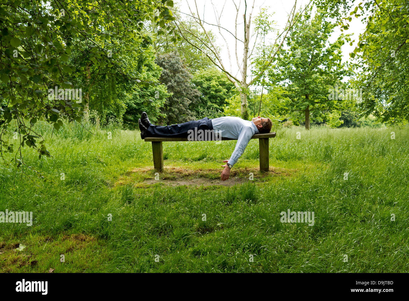 Ein Geschäftsmann liegt in Rückenlage auf einer Holzbank im Regents Park, London. Landschaft. Stockfoto