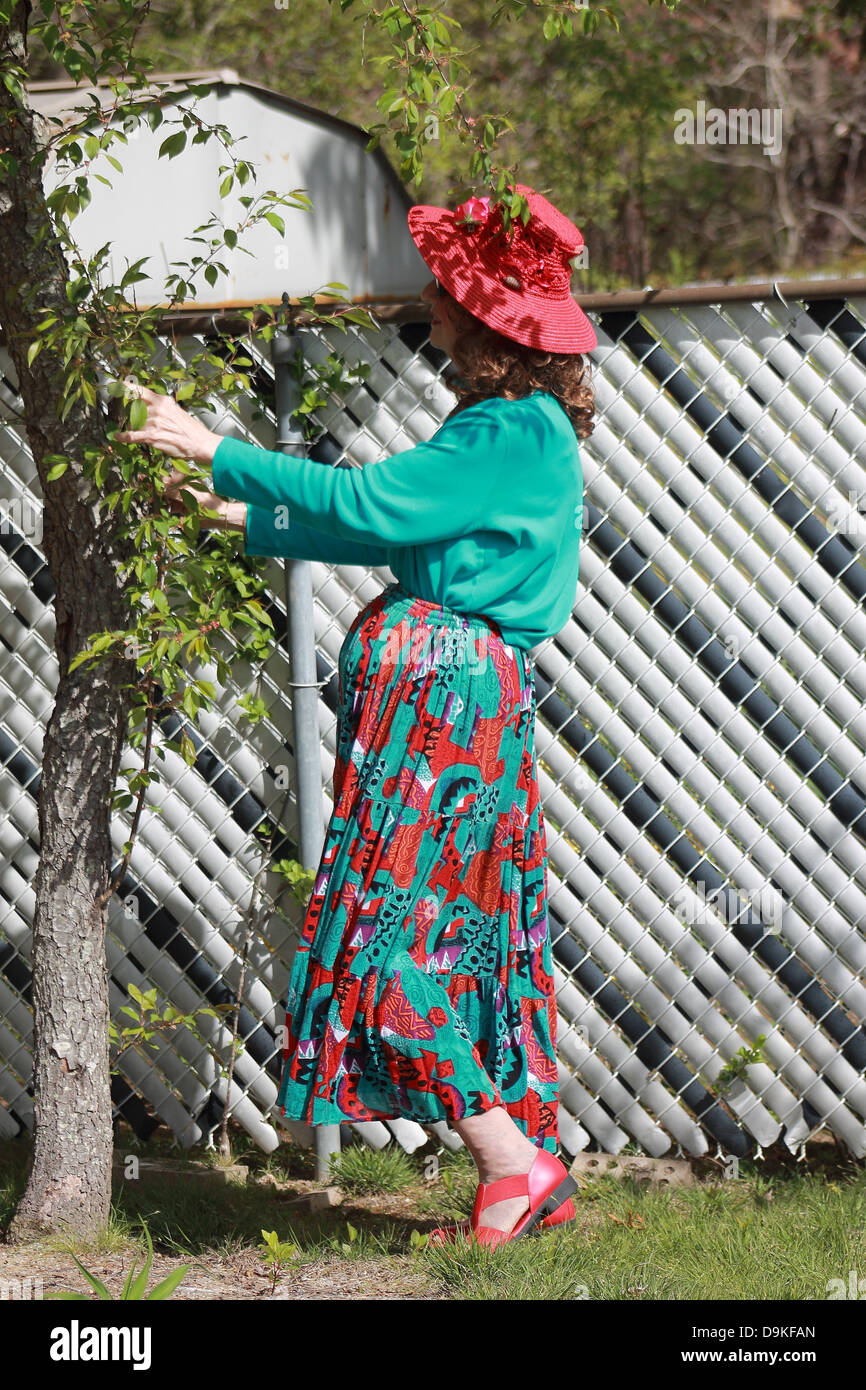 Frau in einem bunten Kleid und roten Hut Beschneiden eines Baumes im freien Stockfoto