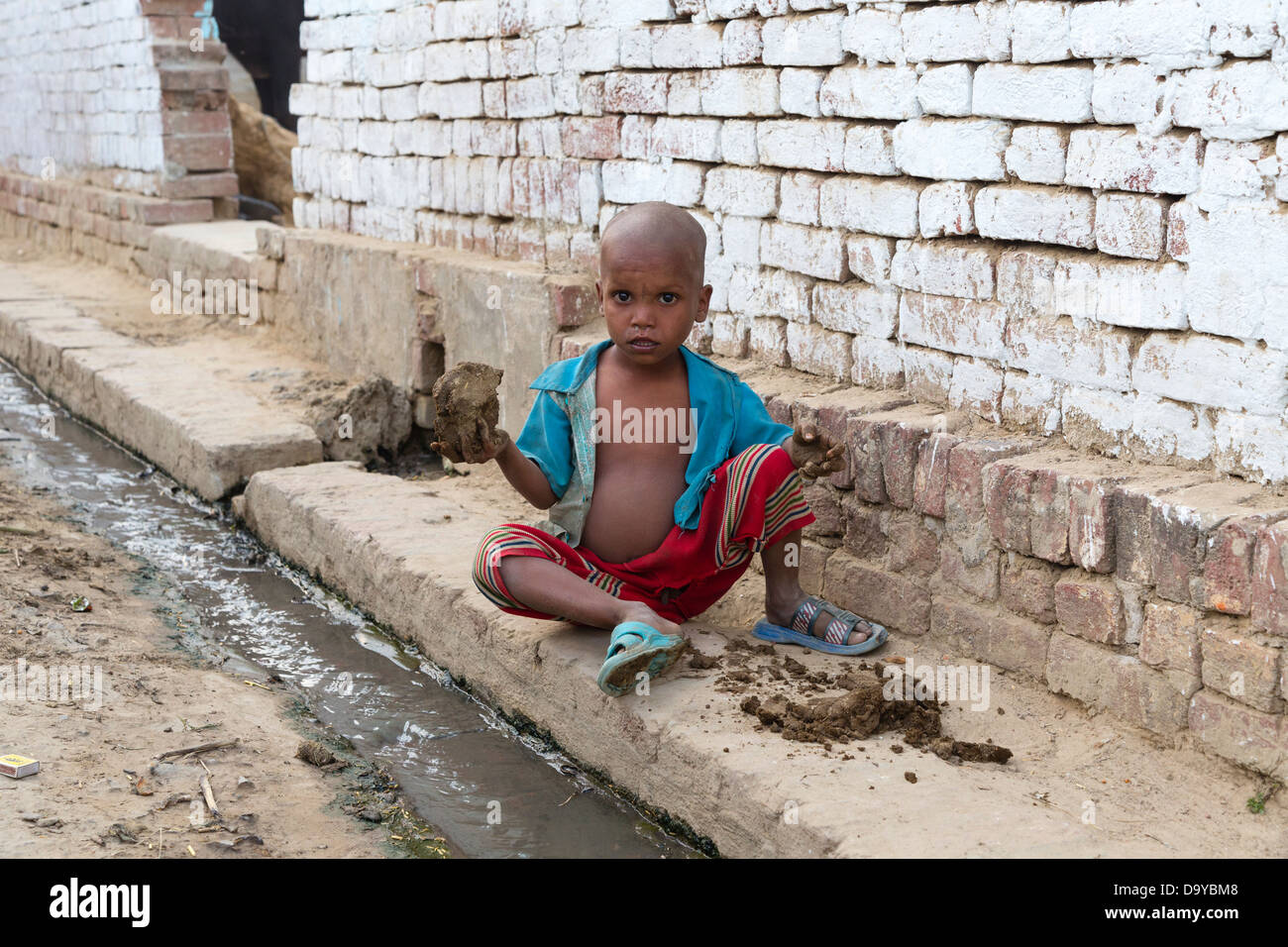 Indien, Uttar Pradesh, Aligarh, sehr kleiner Junge spielt mit Kuhdung neben open-drain Stockfoto