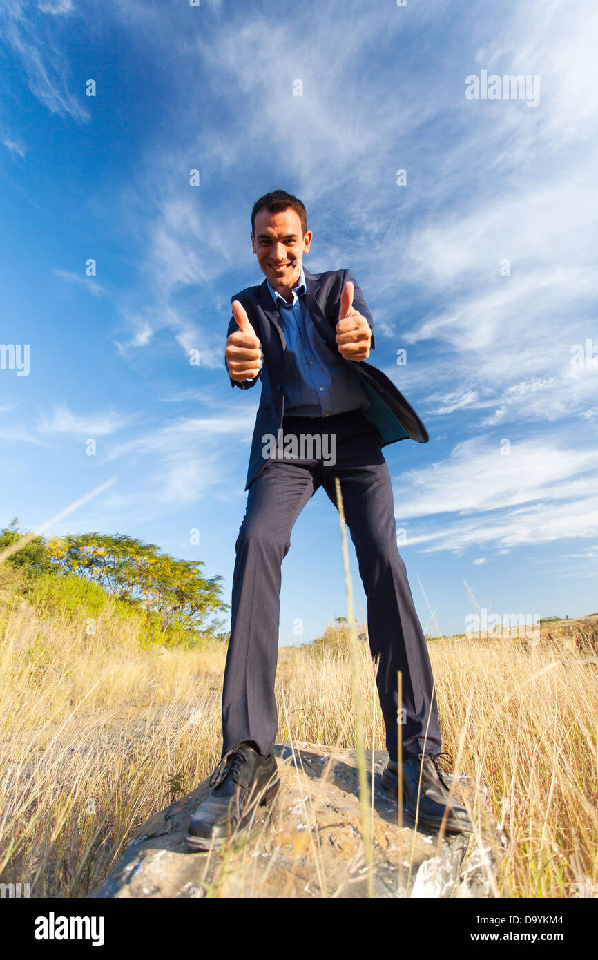 fröhliche junge Geschäftsmann auf Felsen geben Daumen nach oben im freien Stockfoto