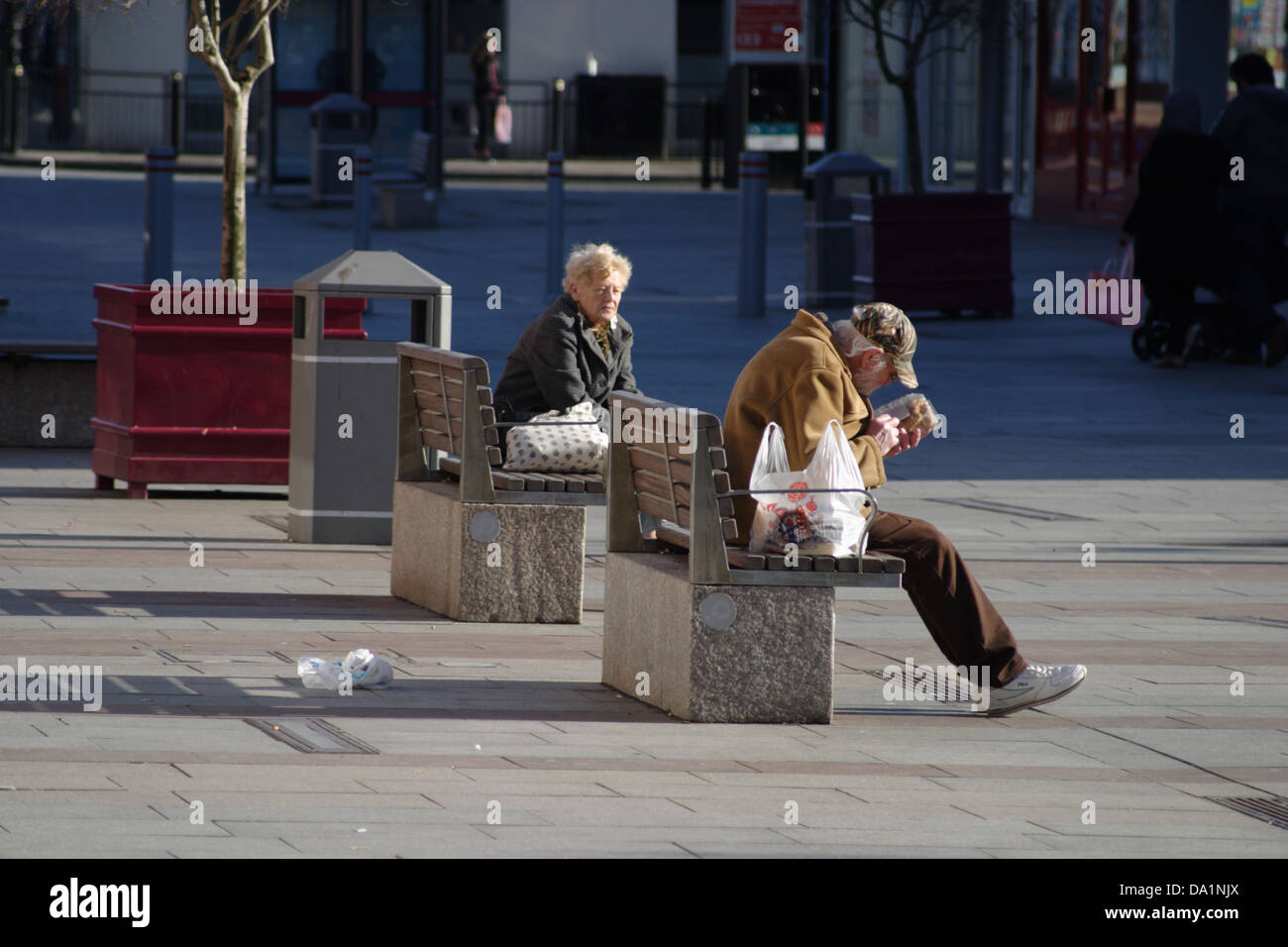 Mann und Frau sitzen auf Bänken im Stadtzentrum trennen. Man öffnet ein Paket von Lebensmitteln, hat eine Tragetasche. Frau hat Handtasche. Stockfoto