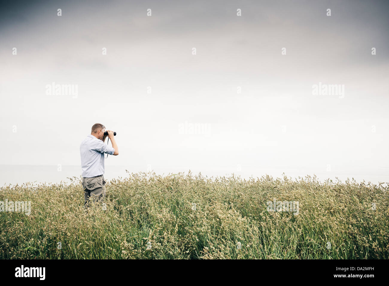 Mann in langen Gräsern auf einer Klippe in Newquay, Cornwall, Blick auf das Meer mit einem Fernglas. Blick in die Zukunft Stockfoto