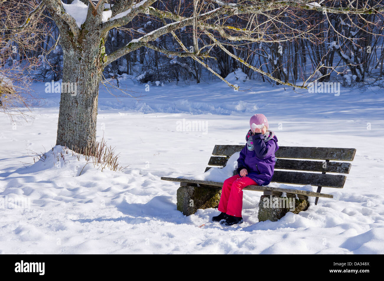 Kleines Mädchen auf einer Bank unter einem Baum im verschneiten Umgebung Stockfoto