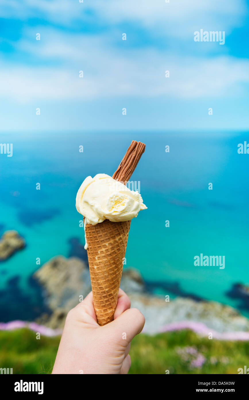 Hand, die Eiswaffel in Newquay, Cornwall, England, gegen das strahlend blaue Meer und den Himmel. Stockfoto
