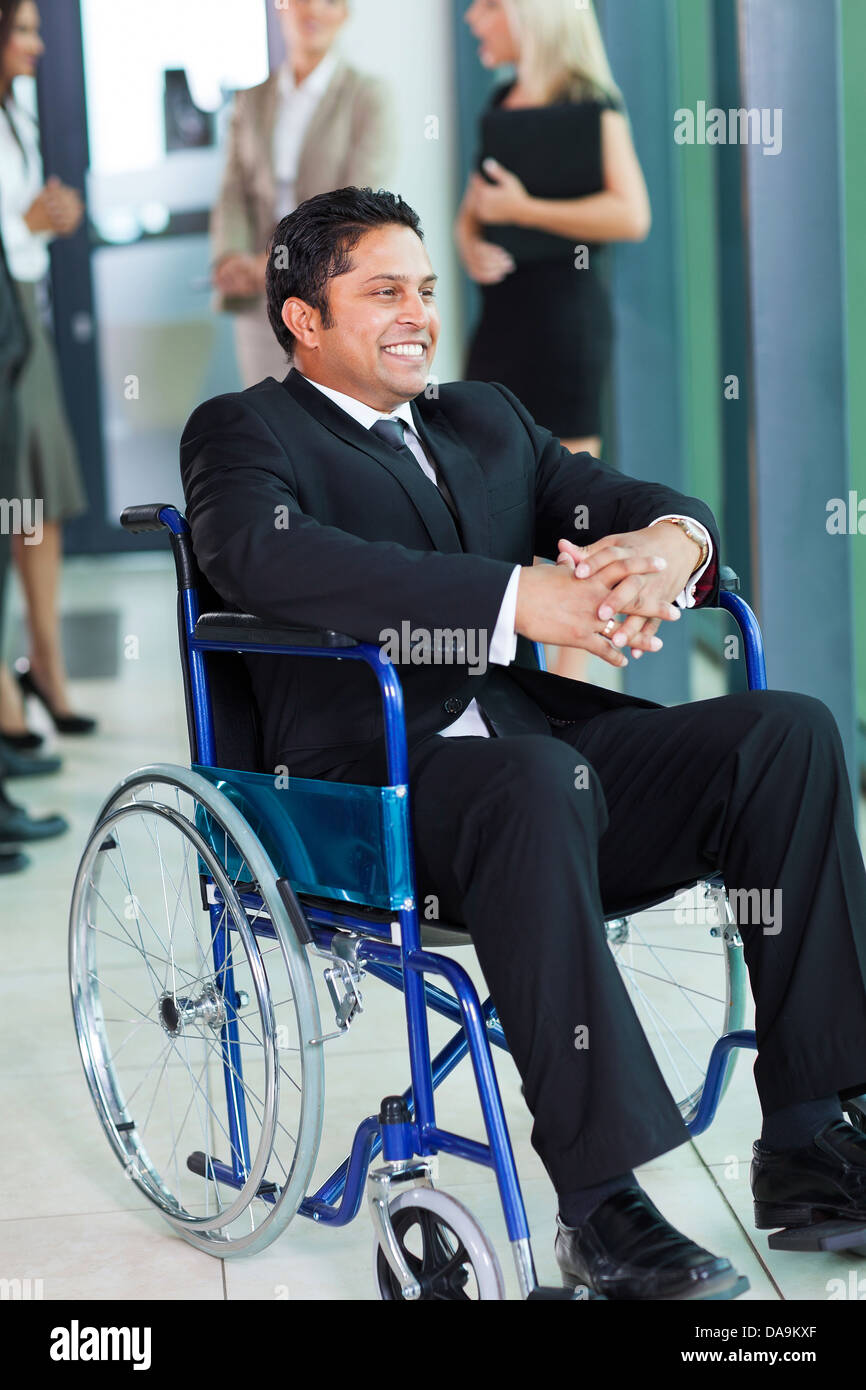 fröhliche indischer Geschäftsmann im Rollstuhl mit Kolleginnen und Kollegen im Hintergrund Stockfoto