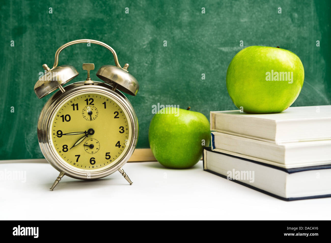 Schule mal wieder. Uhr, Äpfel und Bücher über Lehrer Tabelle, grüne Tafel im Hintergrund. Zurück zur Schule. Stockfoto