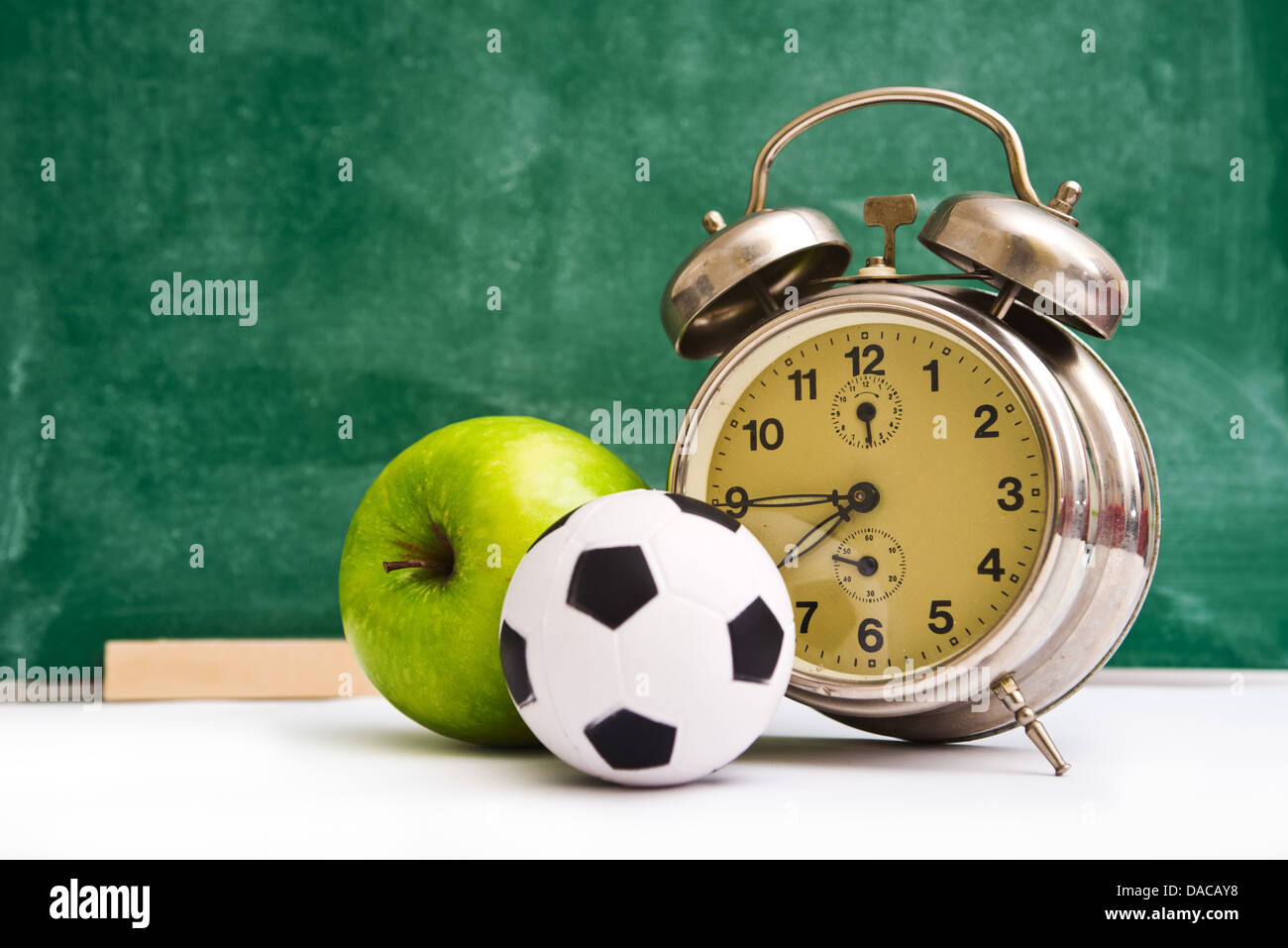 Schule mal wieder. Uhr, kleine Kugel und Apfel an Lehrer Tabelle, grüne Tafel im Hintergrund. Zurück zur Schule. Stockfoto