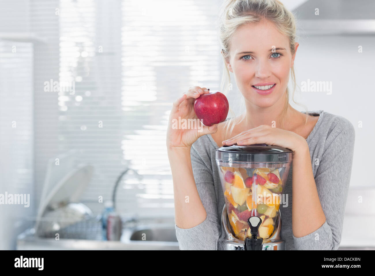Glückliche Frau stützte sich auf ihre Saftpresse voller Früchte und hält roten Apfel Stockfoto