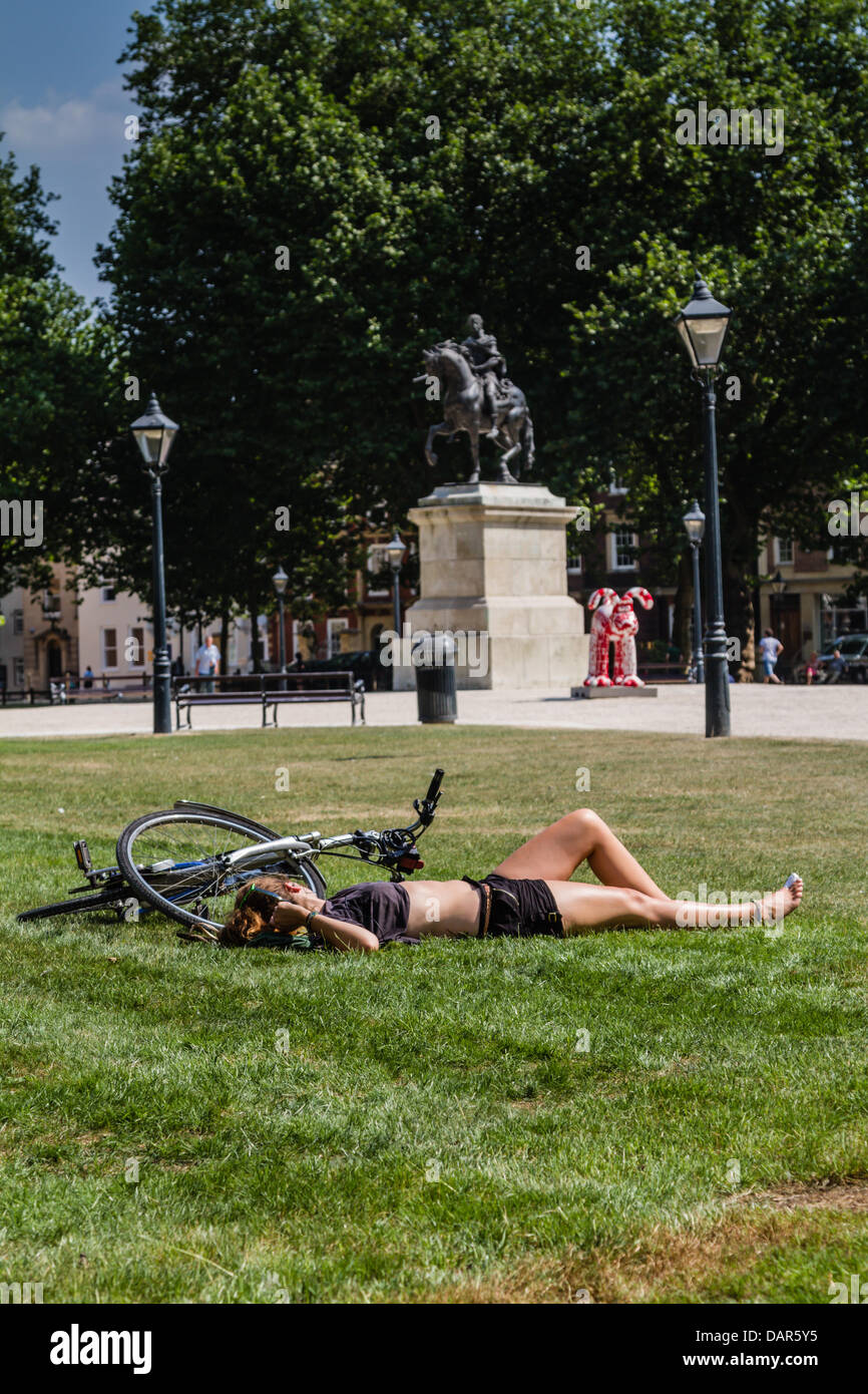 Bristol, UK. 17. Juli 2013. Ein weibliche Radfahrer legt auf dem Rasen neben ihr Fahrrad im Queen Square, Bristol in der Sonne. Gromit Statue sehen Sie im Hintergrund, Gromit Unleashed Kunst Spazierstrecke Credit: Rob Hawkins/Alamy Live News Stockfoto