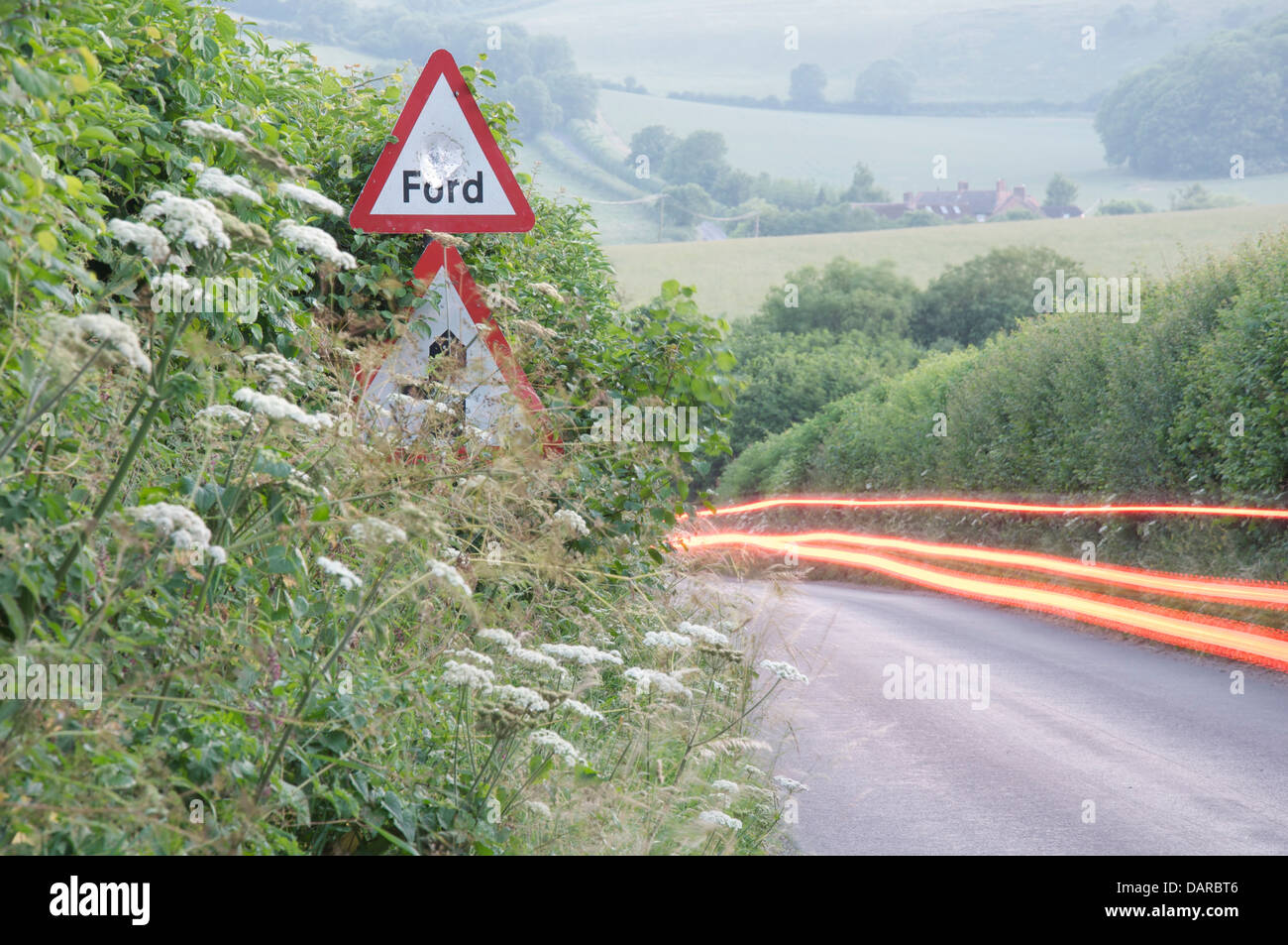 Englische Landschaft. Die roten Rückleuchten eines Auto-Streifen durch die grüne Hecken einen schmalen Feldweg in Dorset. Ein Schild warnt vor einer Furt. Stockfoto