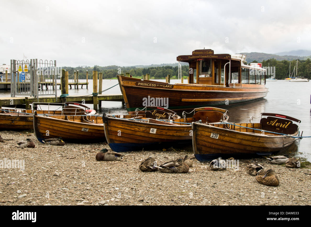 Einige Ruderboote und ein Fahrgastschiff am Ufer des Lake Windermere neben hölzerne Pier. Graue Wolken überhängenden entfernten Hügel. Stockfoto