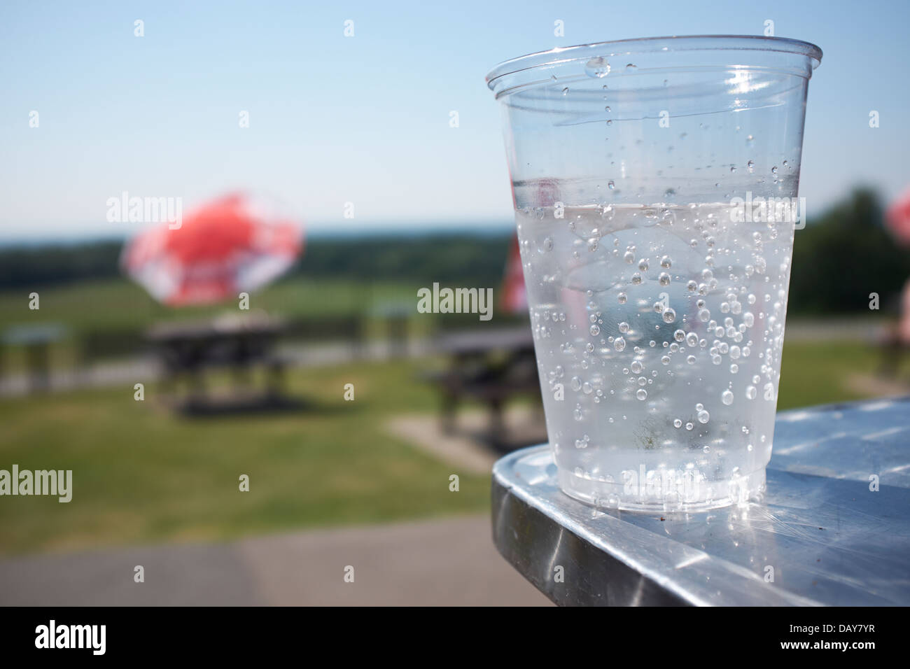 Ein Plastikbecher kohlensäurehaltiges Wasser auf einem Tisch in einem Park-Café mit grünem Rasen und blauen Himmel Hintergrund kurz Schärfentiefe Stockfoto