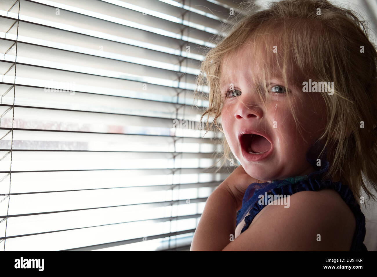 Eine blonde weibliche kaukasischen Säugling weint & betrübt über ein Fenster bedeckt mit Jalousien Stockfoto