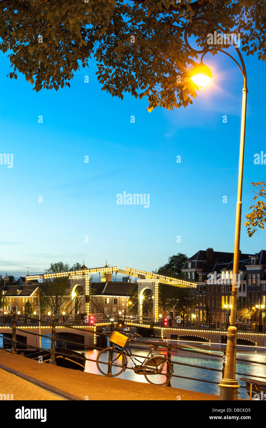 Amsterdam Magere Brug Skinny Bridge am Fluss Amstel Dämmerung Nacht Abend im Sommer. Ein einzelnes Fahrrad ist Geländer gesperrt. Stockfoto