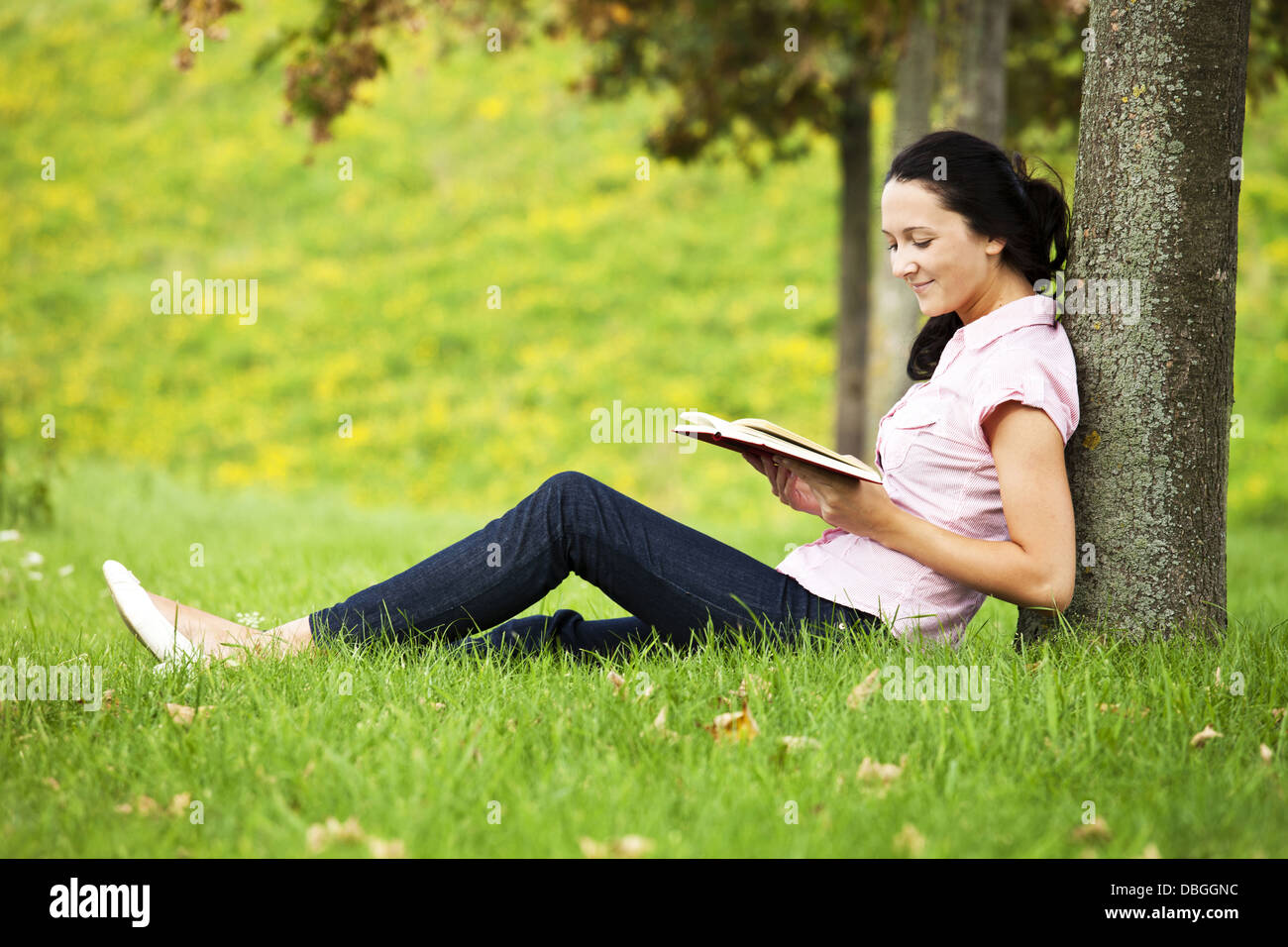 Junge dunkelhaarige Mädchen an einen Baum gelehnt und ein Buch zu lesen Stockfoto