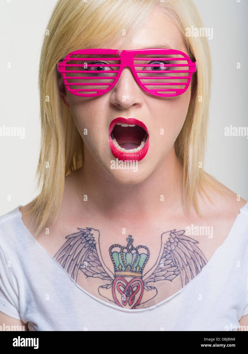 Junge, blonde Kaukasische Frau mit großen Claddagh-Tattoo auf Brust geschlitzte Sonnenbrille rosa Stockfoto