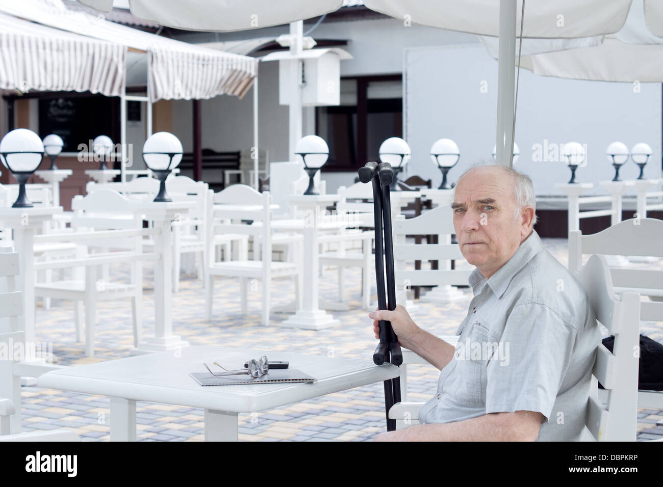 Älterer Mann sitzt an einem Tisch in einem Restaurant im Freien mit seinen Krücken in der Hand mit einer schweren involviert in die Kamera schaut Stockfoto