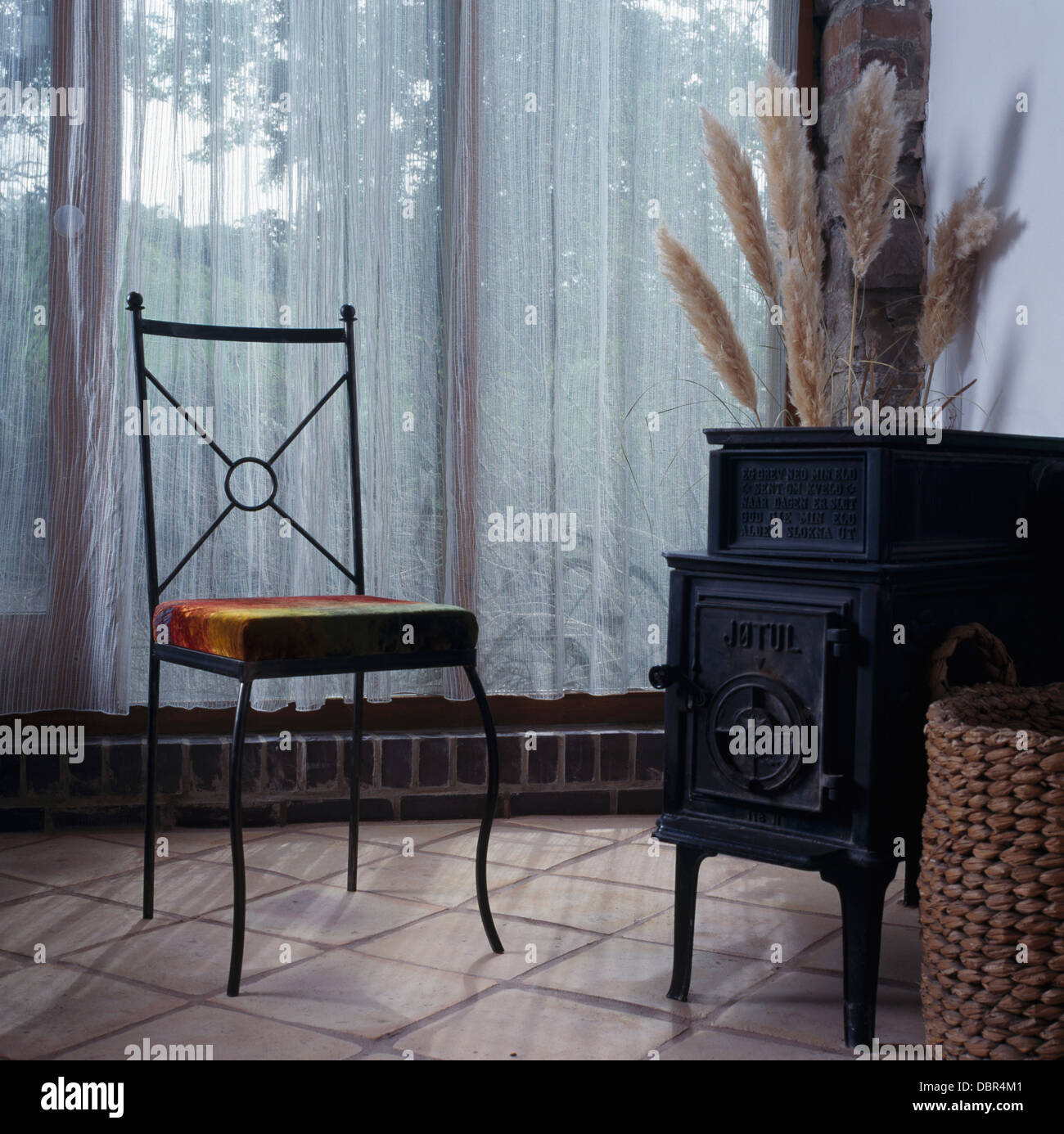 Leder-sitziger Metallstuhl neben schwarzen gusseisernen Jotul Kaminofen im Esszimmer mit weißen Voile Vorhänge Stockfoto