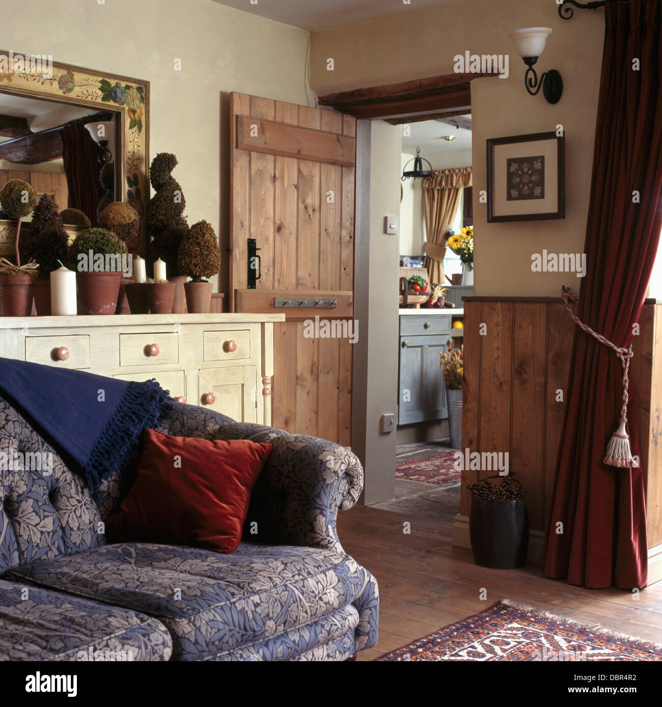 Roter Samt Kissen auf grau-blau floral Sofa im Wohnzimmer Cottage mit abisolierten Kiefer Dado Wandverkleidungen und Kiefer Tür zur Küche Stockfoto