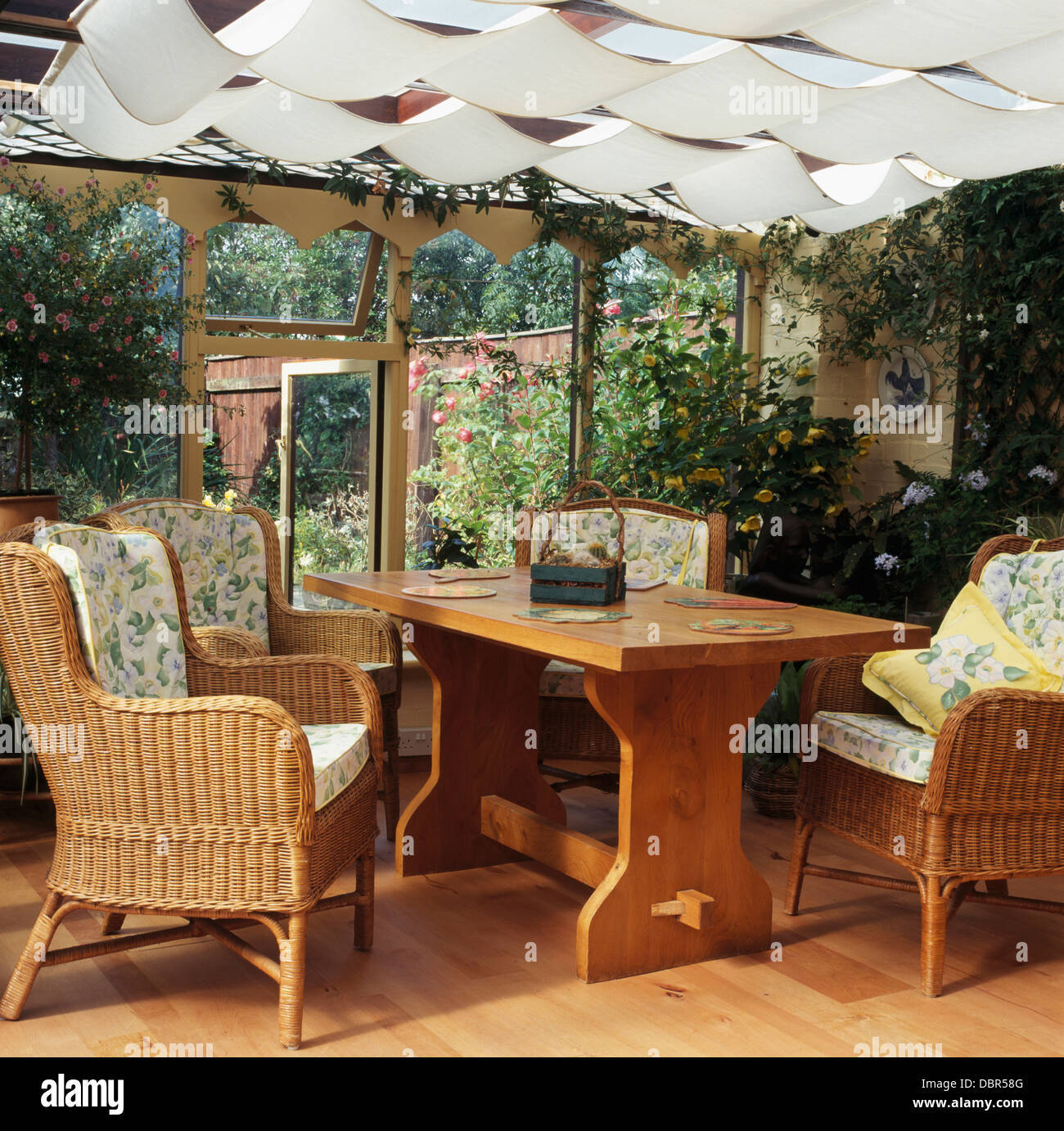 Korbsessel und einfachen Holztisch im Wintergarten mit weißen Decke Jalousien und Blick auf den Garten Stockfoto