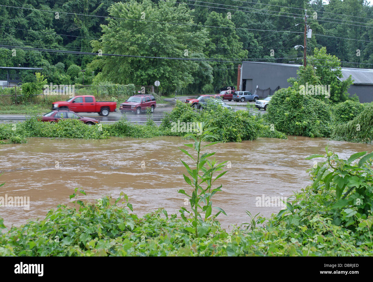 Ein Sturm angeschwollenen Fluss läuft hoch und schlammigen über Überlauf die Ufer und überschwemmen die Autos und die Straße daneben Stockfoto
