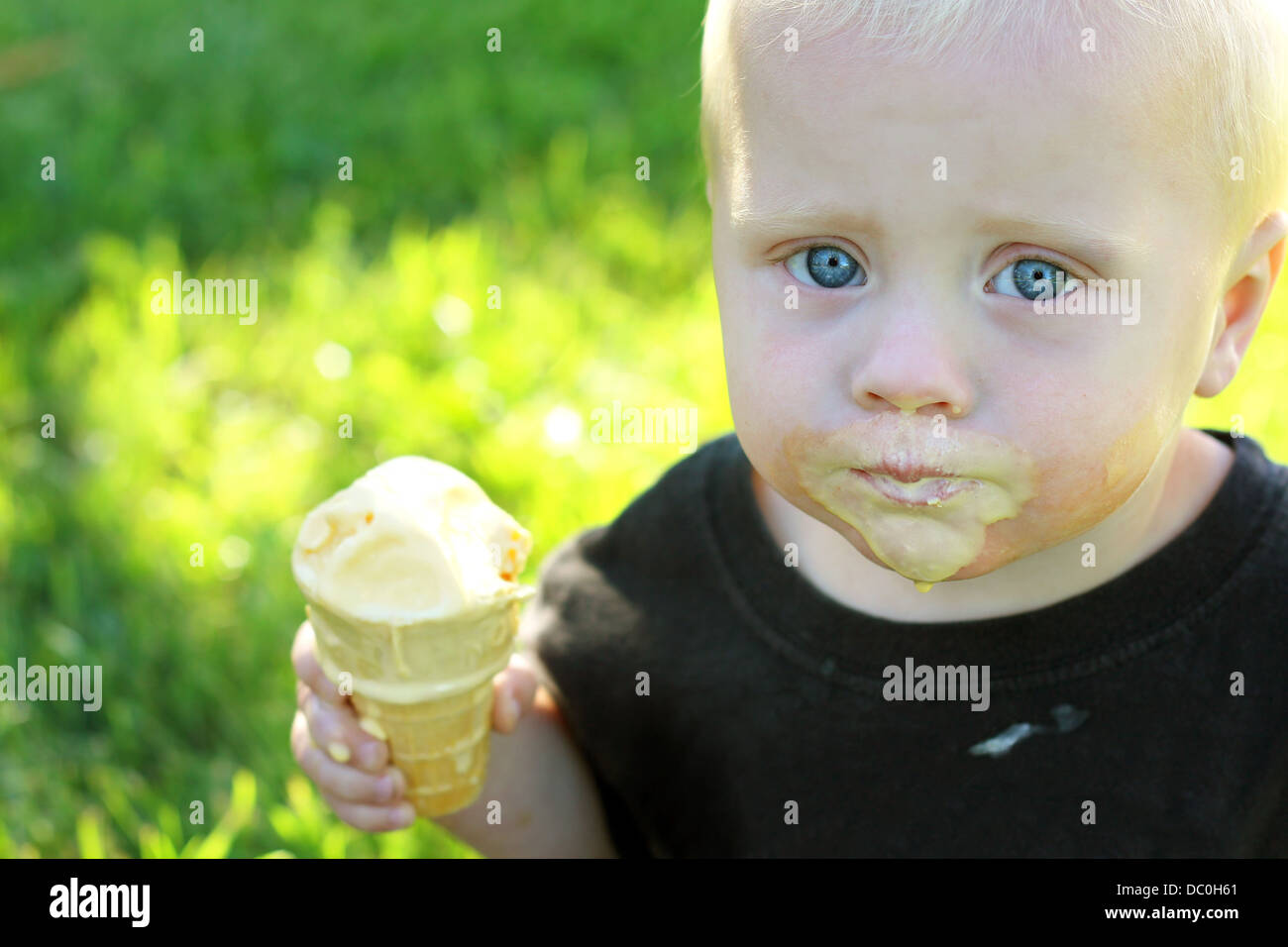ein niedliches Baby Junge sitzt ein unordentlich, schmelzende Vanille Eis essen in den Rasen an einem Sommertag außerhalb. Stockfoto