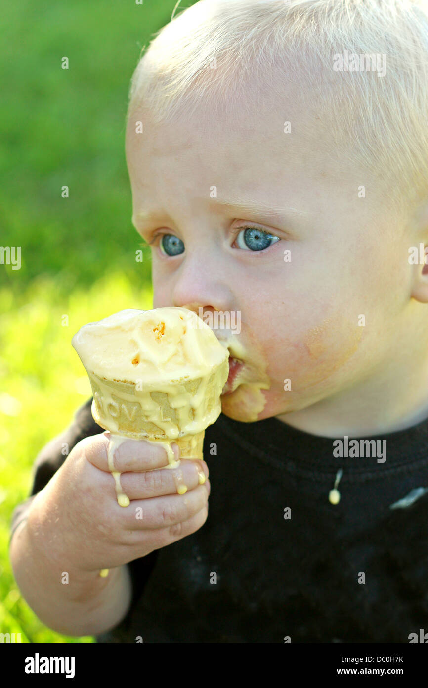 ein süßen, blonden Jungen sitzt außerhalb der Rasen an einem Sommertag in Essen einen Schmelz, chaotisch Vanille Eiscreme-Kegel Stockfoto