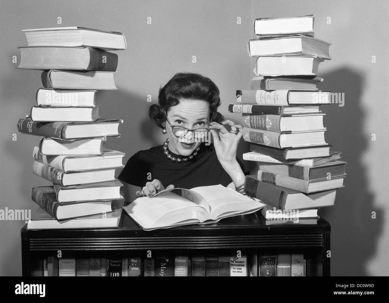 1950ER JAHREN FRAU SITZT ZWISCHEN STAPELN VON BÜCHERN HERUNTERZIEHEN GLÄSER, BLICK ÜBER FELGEN Stockfoto