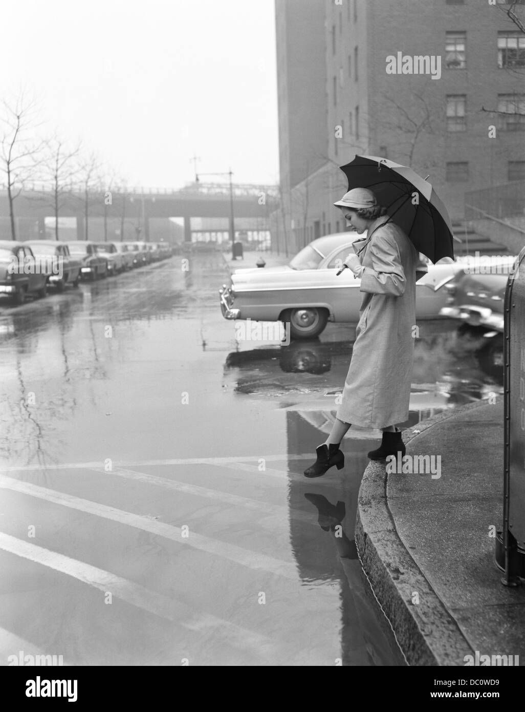 1950ER JAHREN FRAUEN IN MANTEL REGENHUT GUMMISTIEFEL HOLDING-DACH KREUZUNG STADTSTRAßE IN SCHLECHTEM REGENWETTER Stockfoto