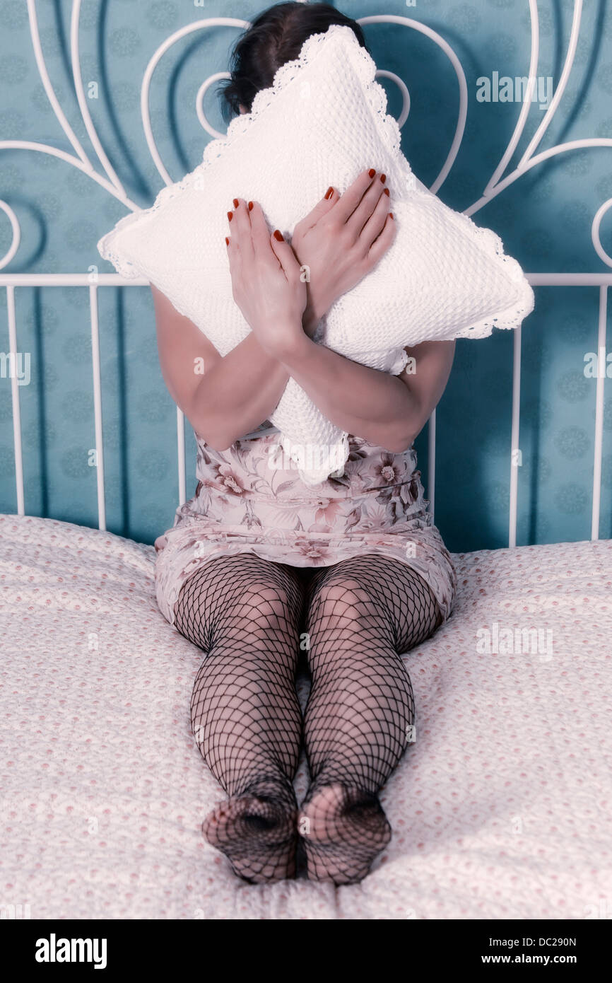 eine Frau in einem Negligé sitzt auf einem Bett, einem Kissen vor ihr Gesicht Stockfoto