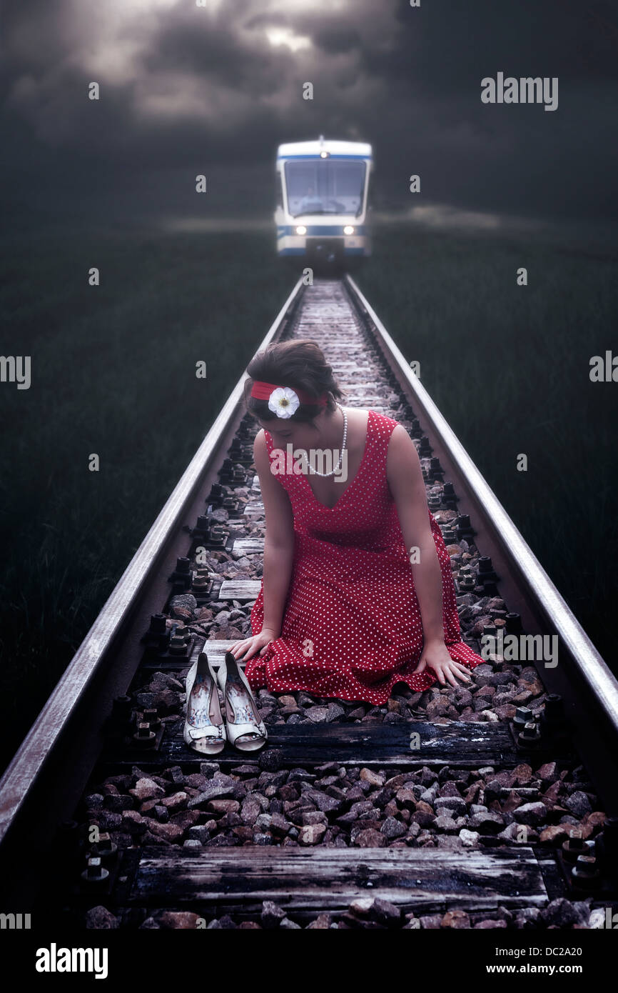 eine Mädchen in einem roten Kleid sitzt auf Schienen, während ein Zug kommt Stockfoto