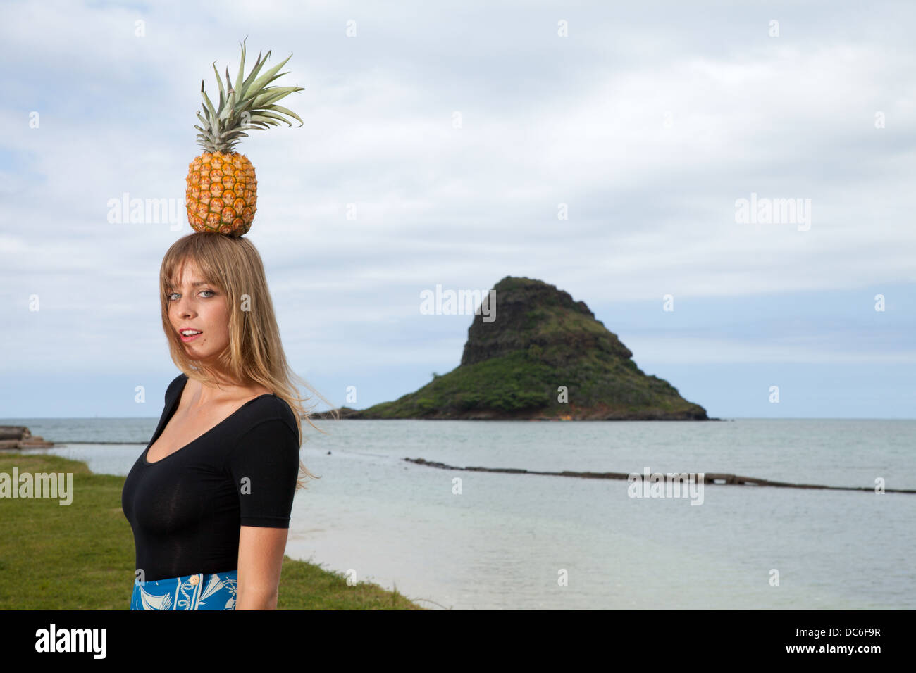 Eine blonde Frau Caucasion steht unter einer Palme mit Ananas auf dem Kopf in der Nähe einer hawaiianischen Insel. Stockfoto
