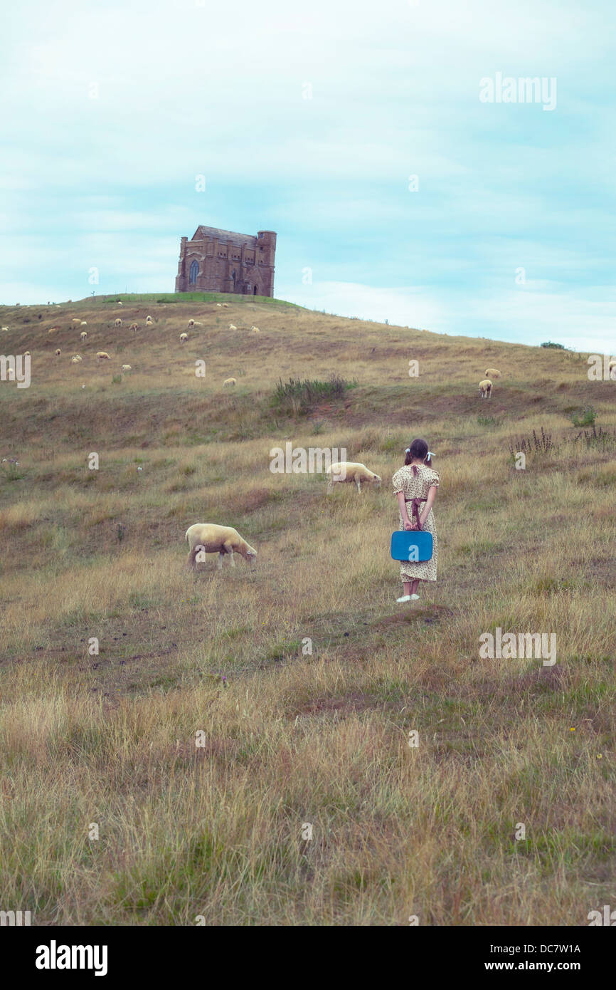 ein Mädchen in ein Vintage-Kleid auf einer Wiese mit Schafen, im Hintergrund eine Kapelle Stockfoto