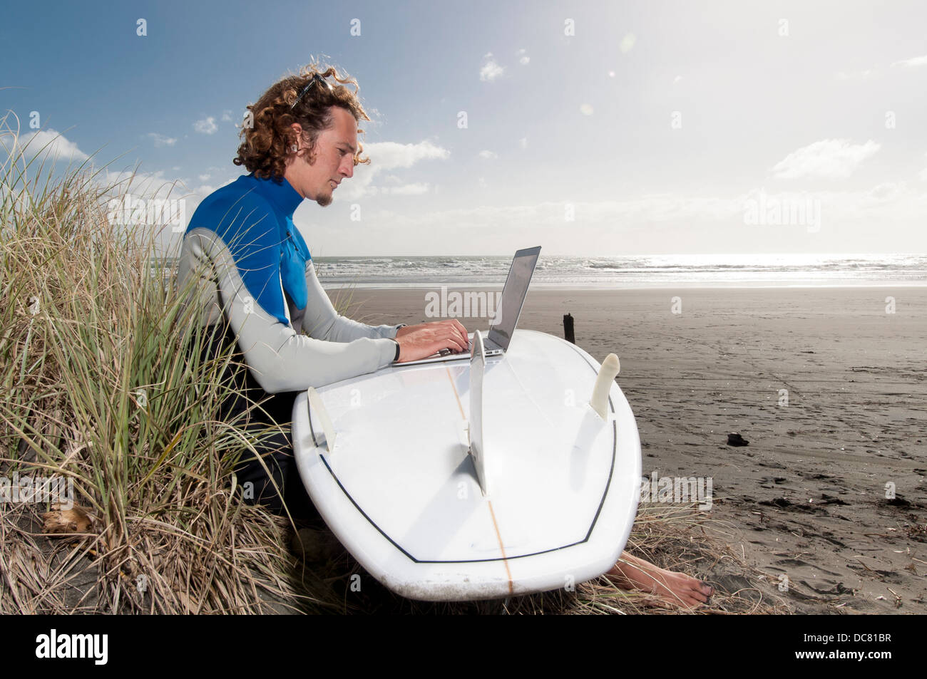 männliche Surfer sitzen am Strand mit Laptop auf Surfbrett, Ngarunui Strand, Raglan, Neuseeland Stockfoto