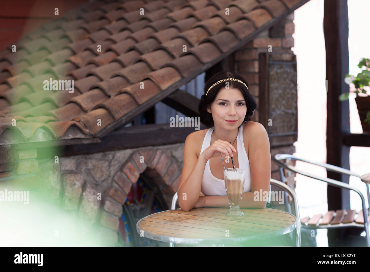 Portrait von schönen jungen Mädchen in einem Sommer Restaurant im Freien mit einem Glas cremigen Cappuccino sitzen. Sie trendige goldene Kette im Haar hat und schönes Lächeln Stockfoto