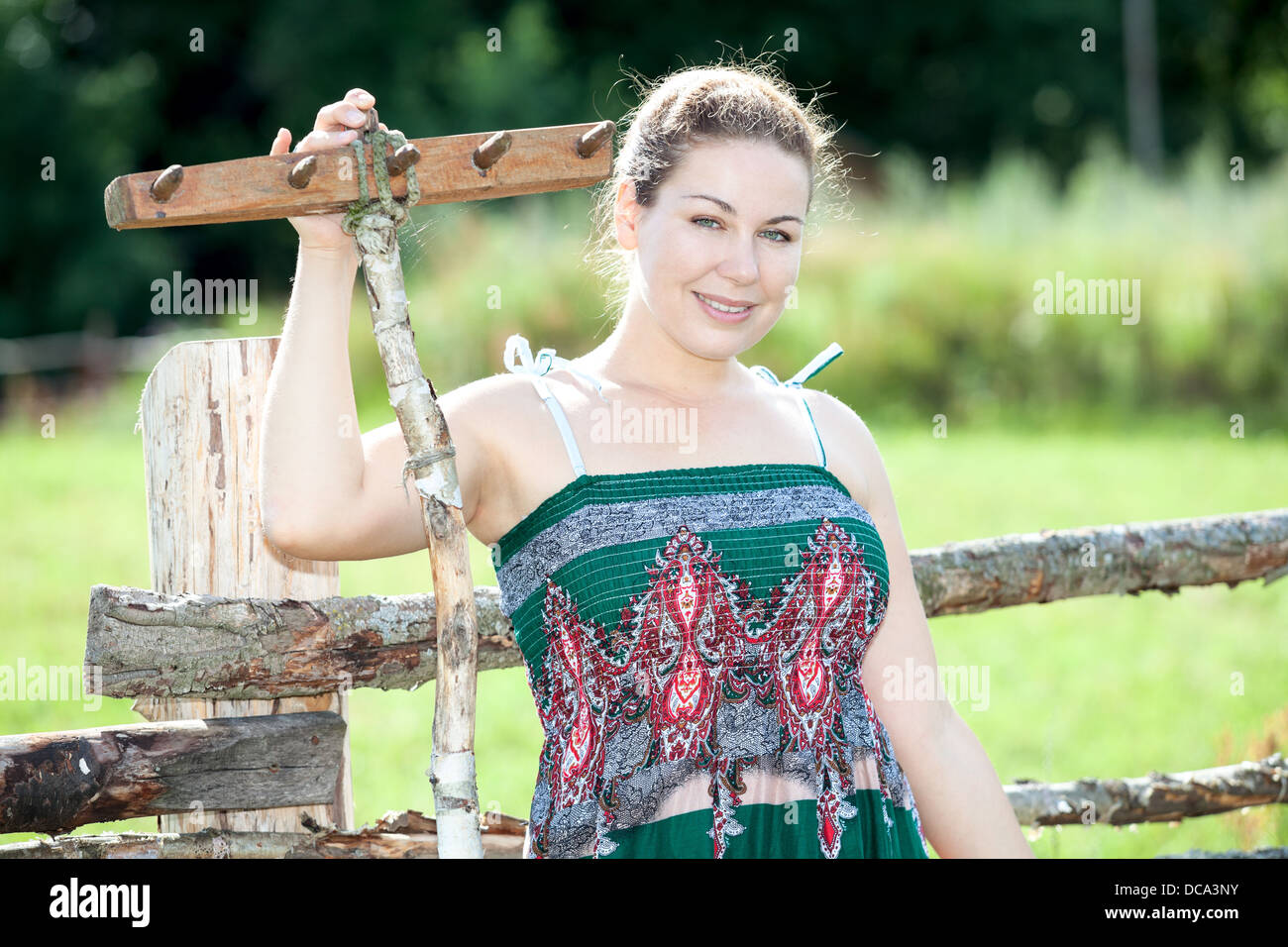 Hübsche Bäuerin stehend mit hölzernen Rechen hinter Zaun im Sommerkleid. Russischen Volkes im ländlichen Russland Stockfoto