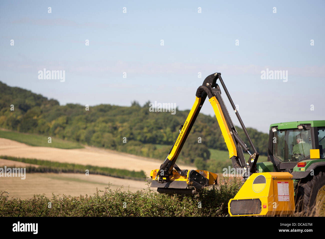 Grün und gelb John Deer Traktor mit Hecke schneiden Arm in Aktion Schneiden einer Hecke am Rande eines Feldes in Oxfordshire, Vereinigtes Königreich. Stockfoto