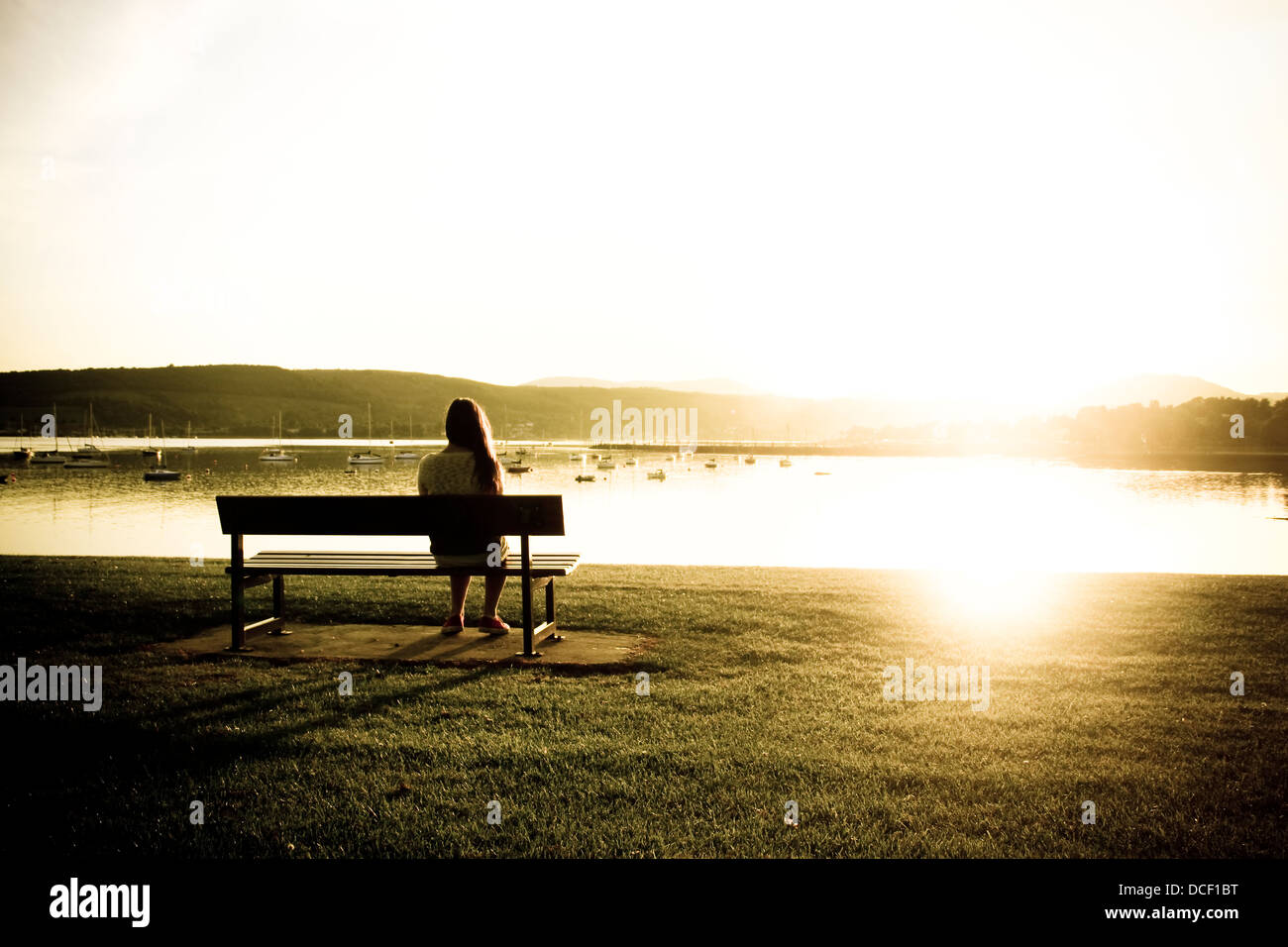 Junges Mädchen auf Parkbank Sonnenuntergang über dem Wasser Stockfoto