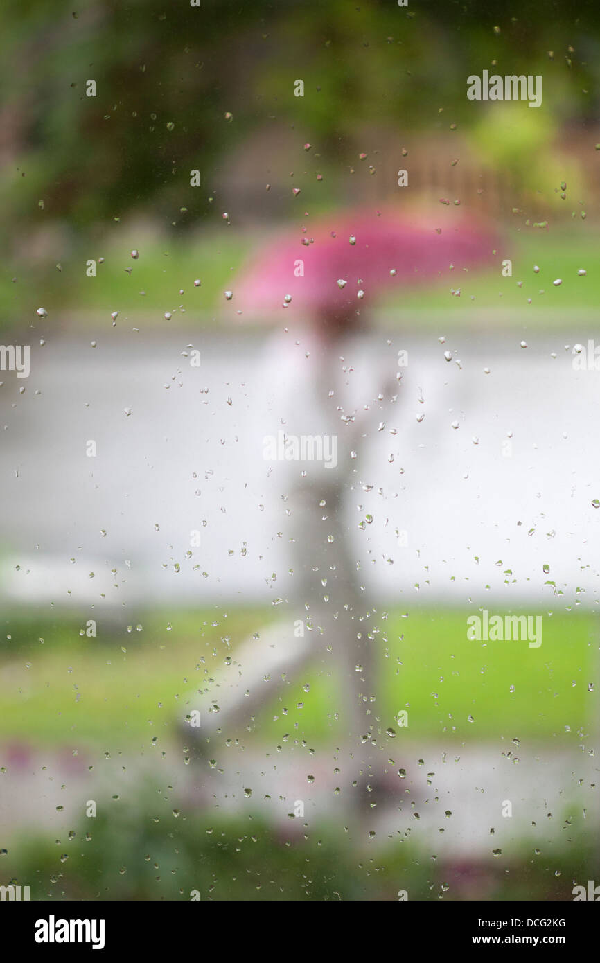 Mann, der einen rosa Regenschirm trägt, der während des Frühjahrsregensturms auf dem Bürgersteig durch Regentropfen auf dem Fensterglas des Hauses läuft Stockfoto