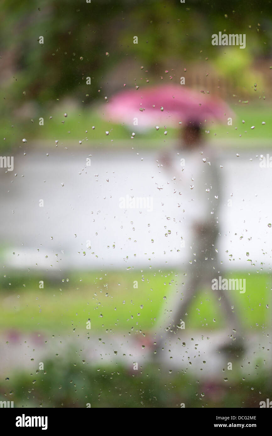Mann, der einen rosa Regenschirm trägt, der während des Frühjahrsregensturms durch Regentropfen auf dem Glasfenster auf dem Bürgersteig läuft Stockfoto