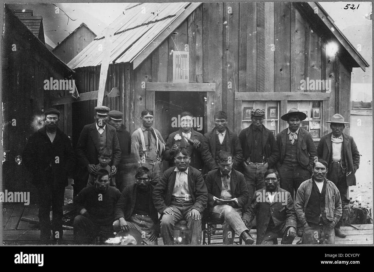 Eine Gruppe von 15 Indianern und einem Jungen versammelte sich vor einem Fotografen-Atelier, aufgenommen in einem historischen Foto, das das kulturelle und gemeinschaftliche Leben während des frühen 20. Jahrhunderts widerspiegelt. Stockfoto