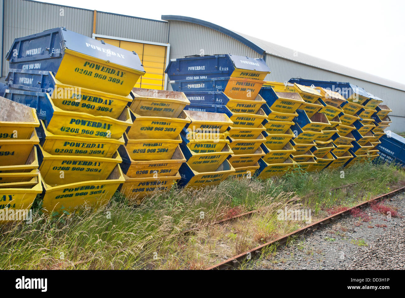 Stapel von stapelten sich gelb und blau Müll "Powerday" überspringt, einige acht tiefen stand in einem Recyclinghof durch eine Eisenbahnlinie in Willesden, Nord-London Stockfoto