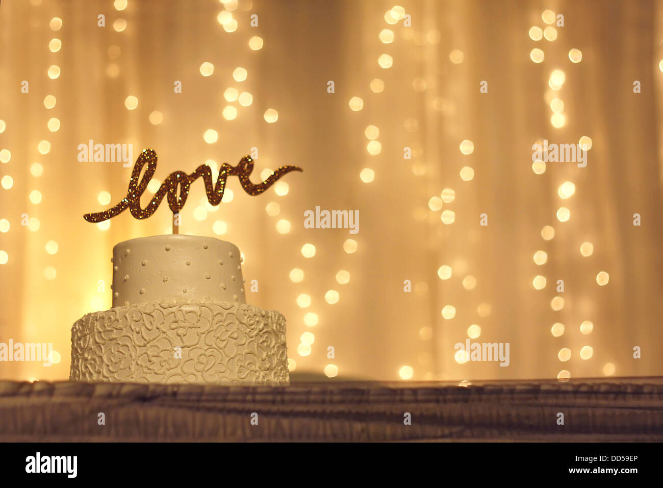 eine einfache weiße Hochzeitstorte mit dem Wort Liebe geschrieben in glitzernden goldenen Lettern auf dem oberen & weißen funkelnden Lichter Hintergrund Stockfoto