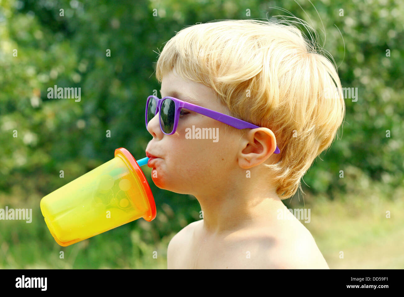ein kleiner Junge mit lila Sonnenbrille ist seinen Kopf zurück, trinken Saft aus einer bunten sippy Tasse an einem sonnigen Sommertag kippen. Stockfoto