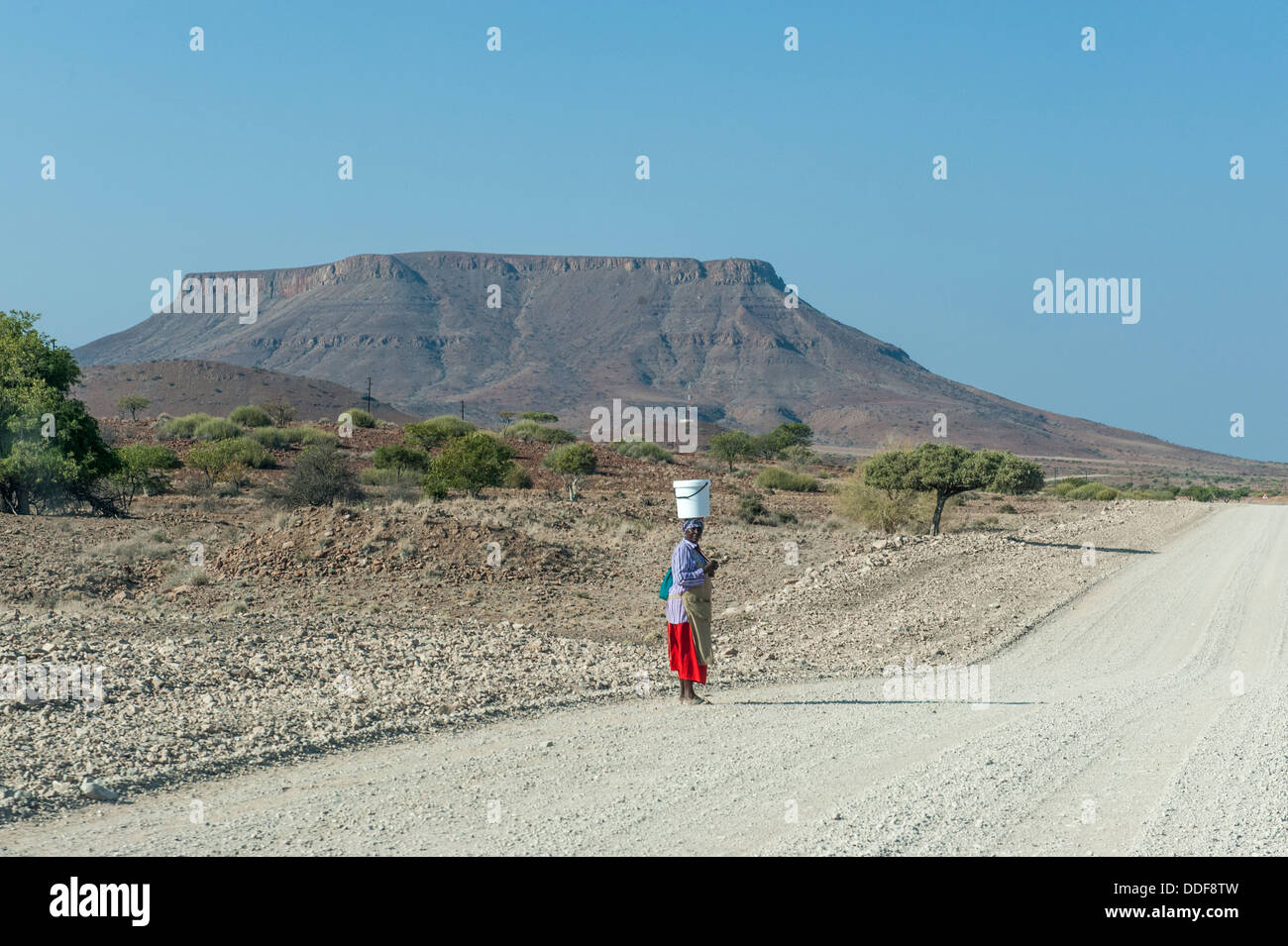 Eine Frau mit einem Eimer auf ihrem Kopf stehend auf einer Schotterstraße in trockene Landschaft, Kunene Region, Namibia Stockfoto