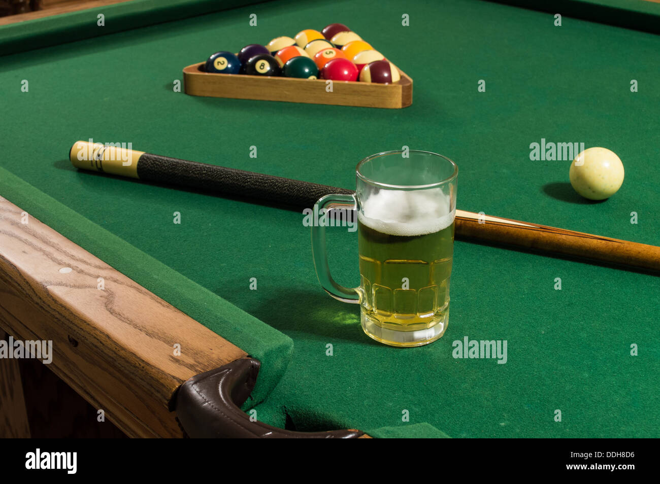 eine kalte Tasse Bier mit Schaum auf einem Billardtisch mit grünem Filz und Rack mit 15 Kugeln, darunter 8-Ball und 9-Ball Pool Stick. Stockfoto