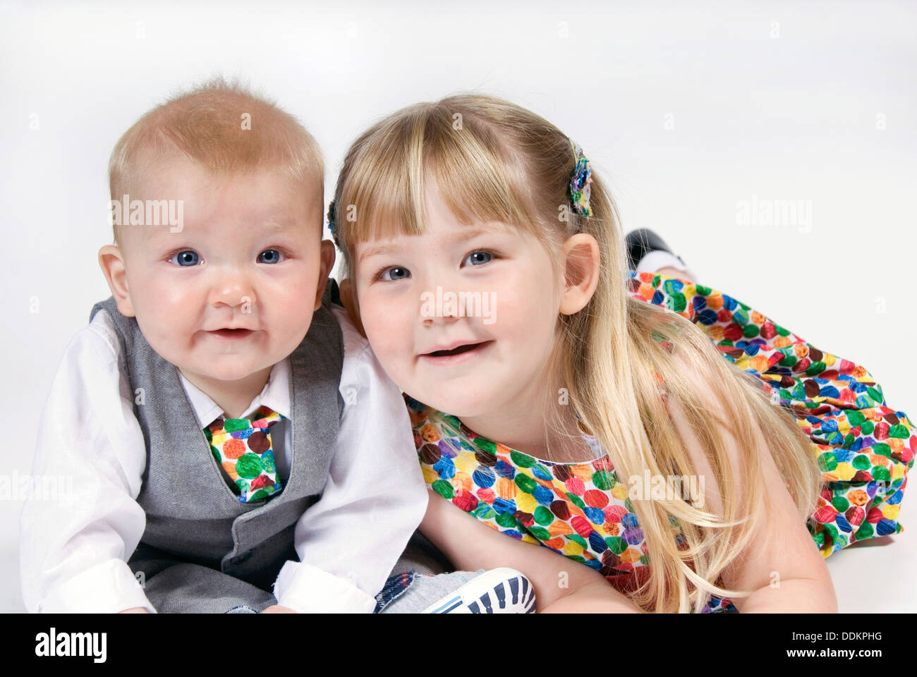 Kleines Mädchen auf ihrem Bauch liegend schmiegt sich zu ihrem Baby Bruder, gekleidet in Anzug und Krawatte und passende nicht so Doll Kleid, UK Stockfoto